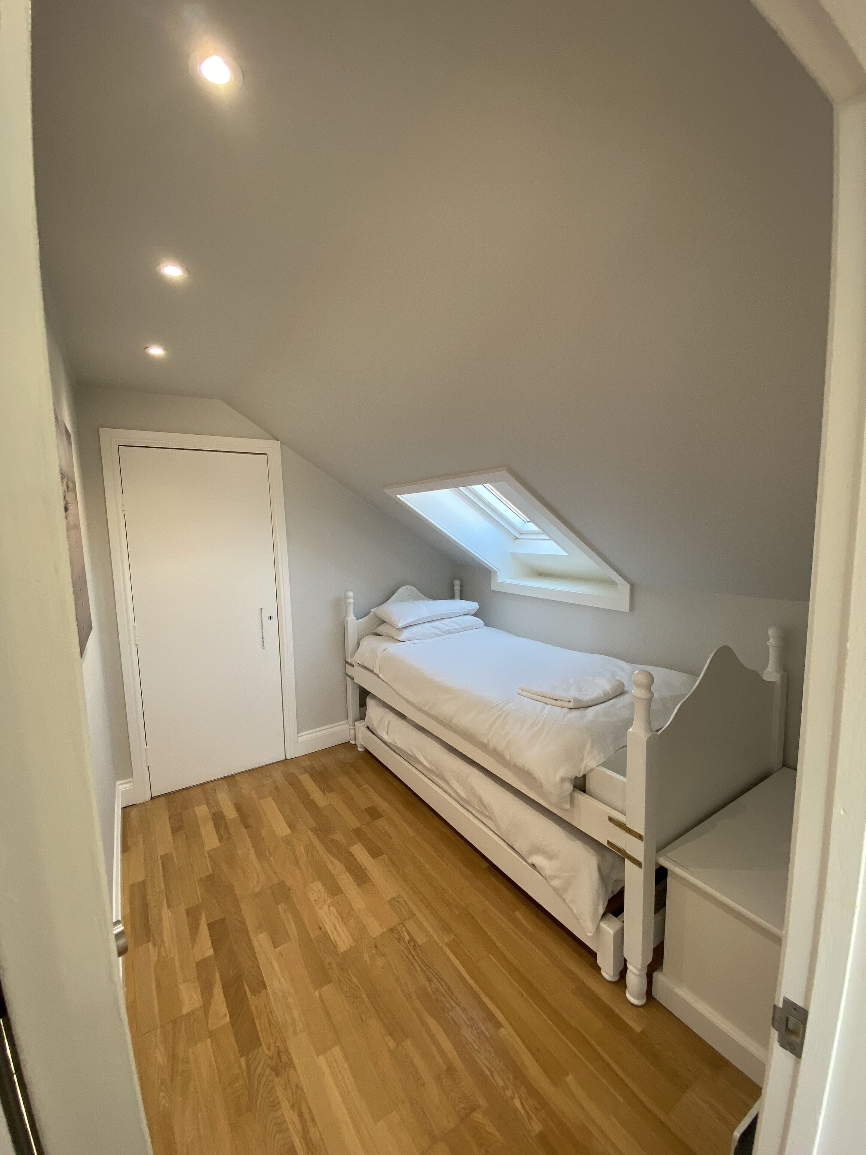 A small, single-bed room with a sloped ceiling and skylight, wooden flooring, and a white door.