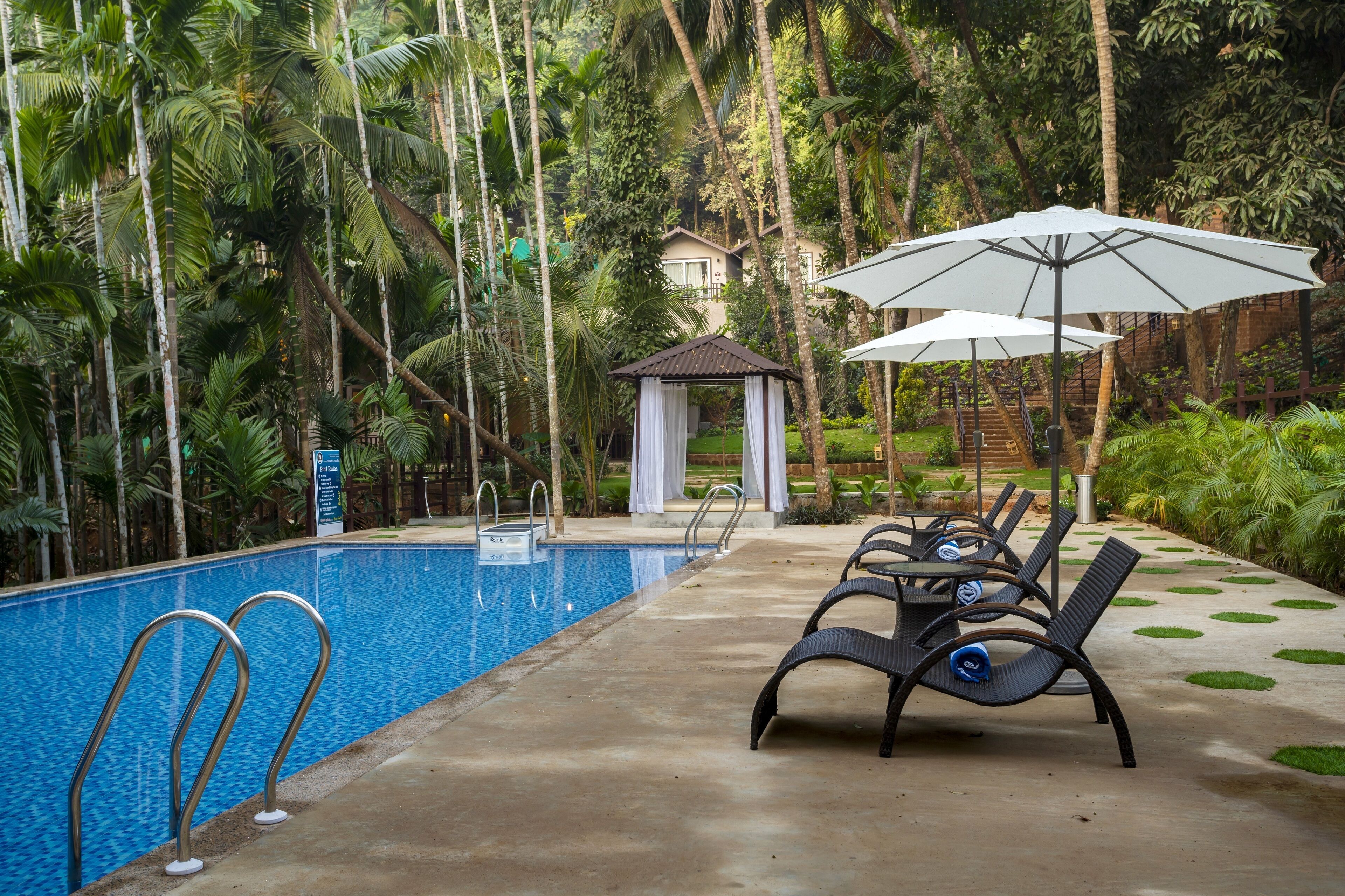 A pool area with lounge chairs and umbrellas, surrounded by lush greenery and a gazebo.