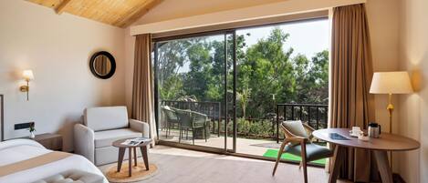 A hotel room with a bed, a sofa, a small table, a chair, a balcony with a view of trees, and a large window.