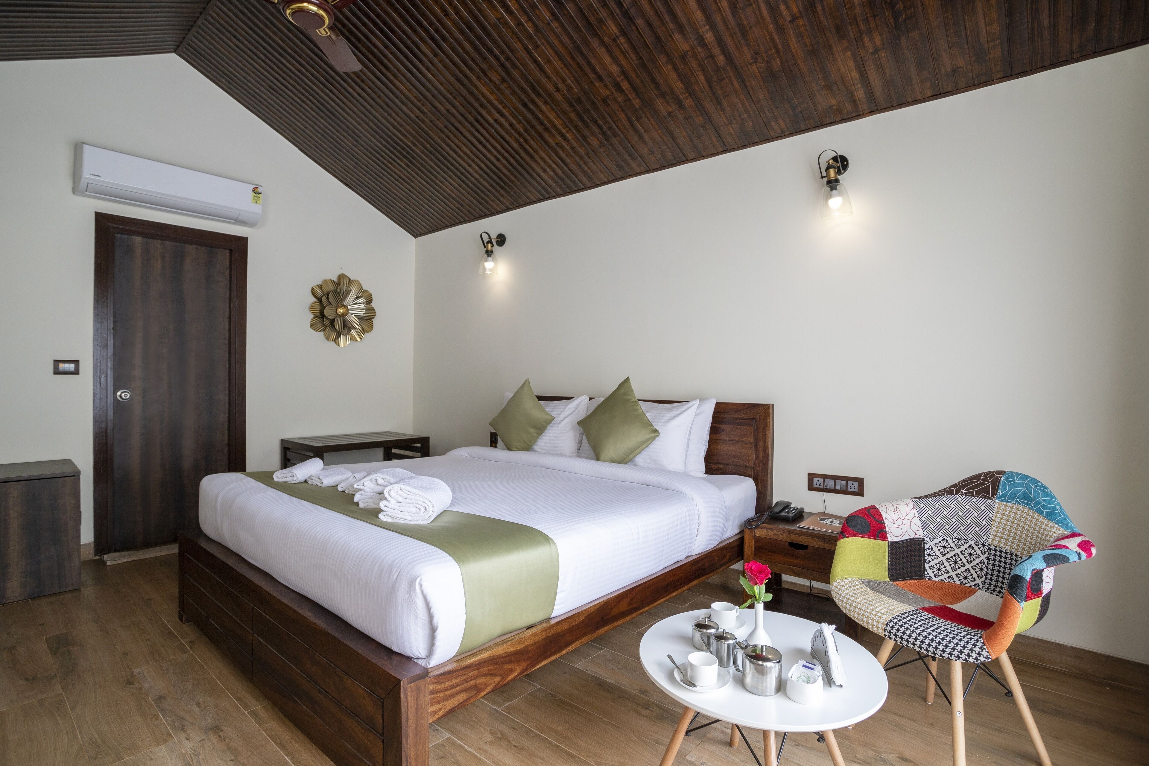 A hotel room with a wooden bed, a round table with a vase of flowers, and a unique patchwork chair.