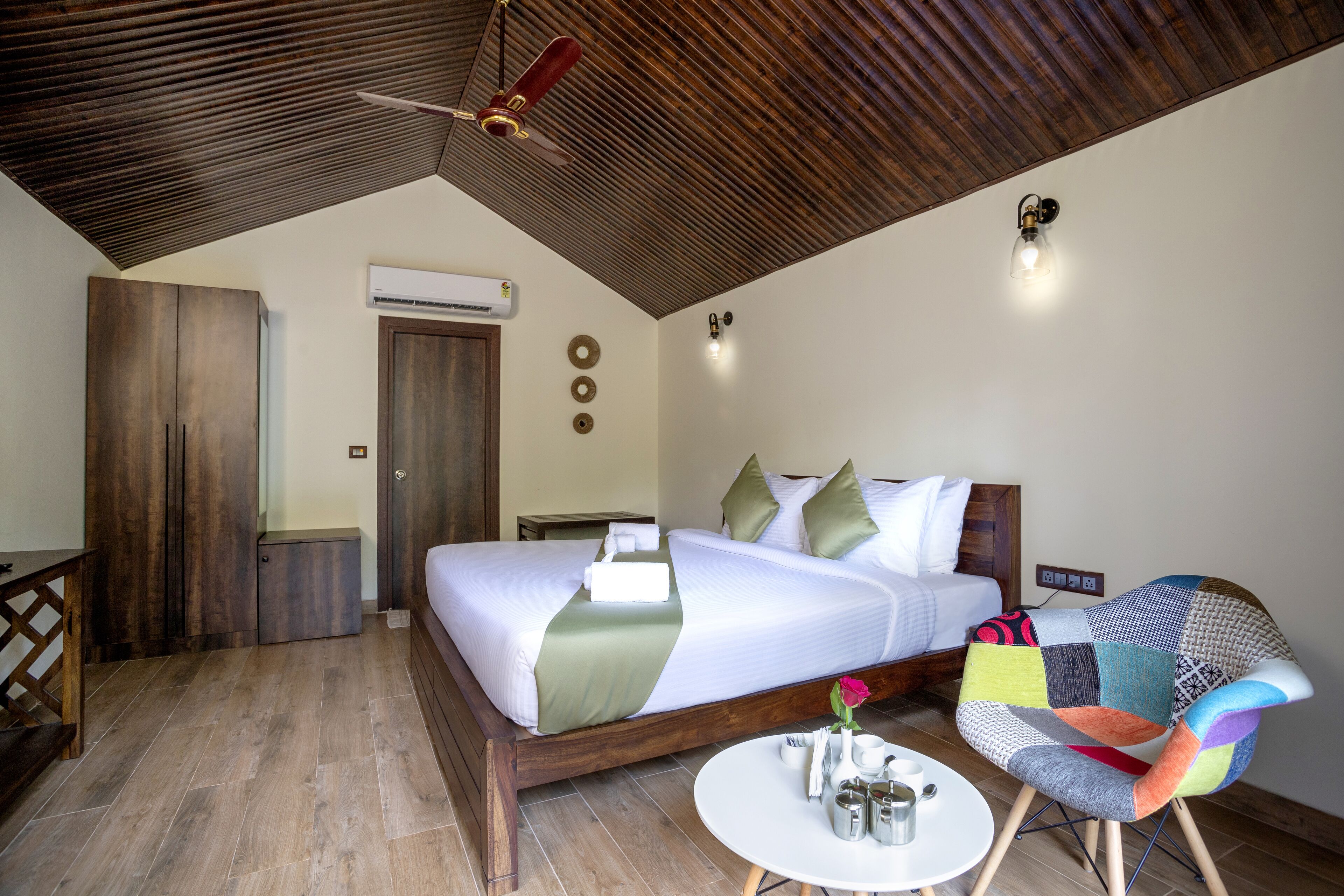 Een slaapkamer met een houten plafond, een bed met kussens, een stoel, een klein tafeltje met kopjes, en een airconditioning.'