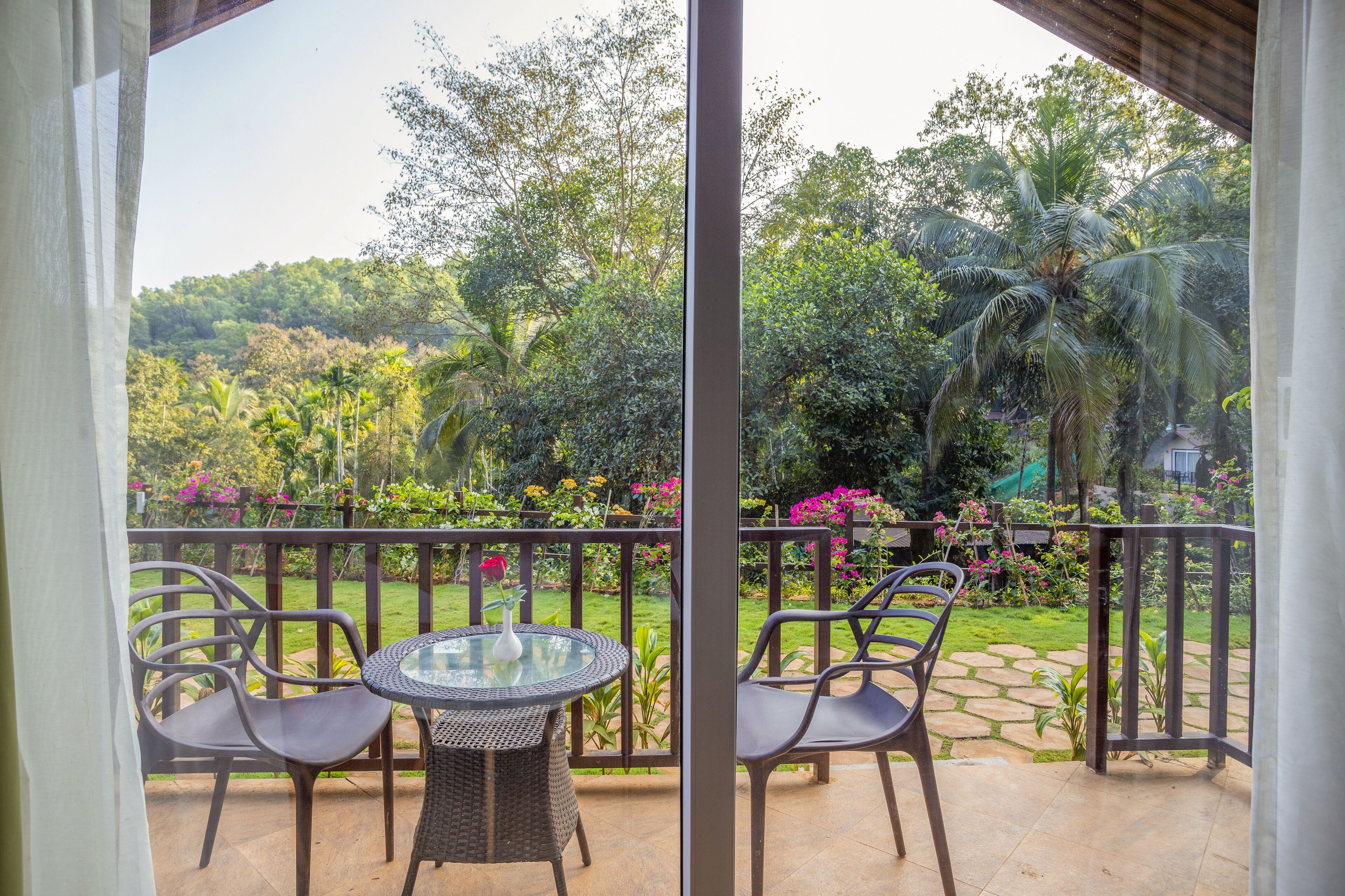 A balcony with wicker chairs and a glass table, overlooking a lush garden with various plants and a palm tree.
