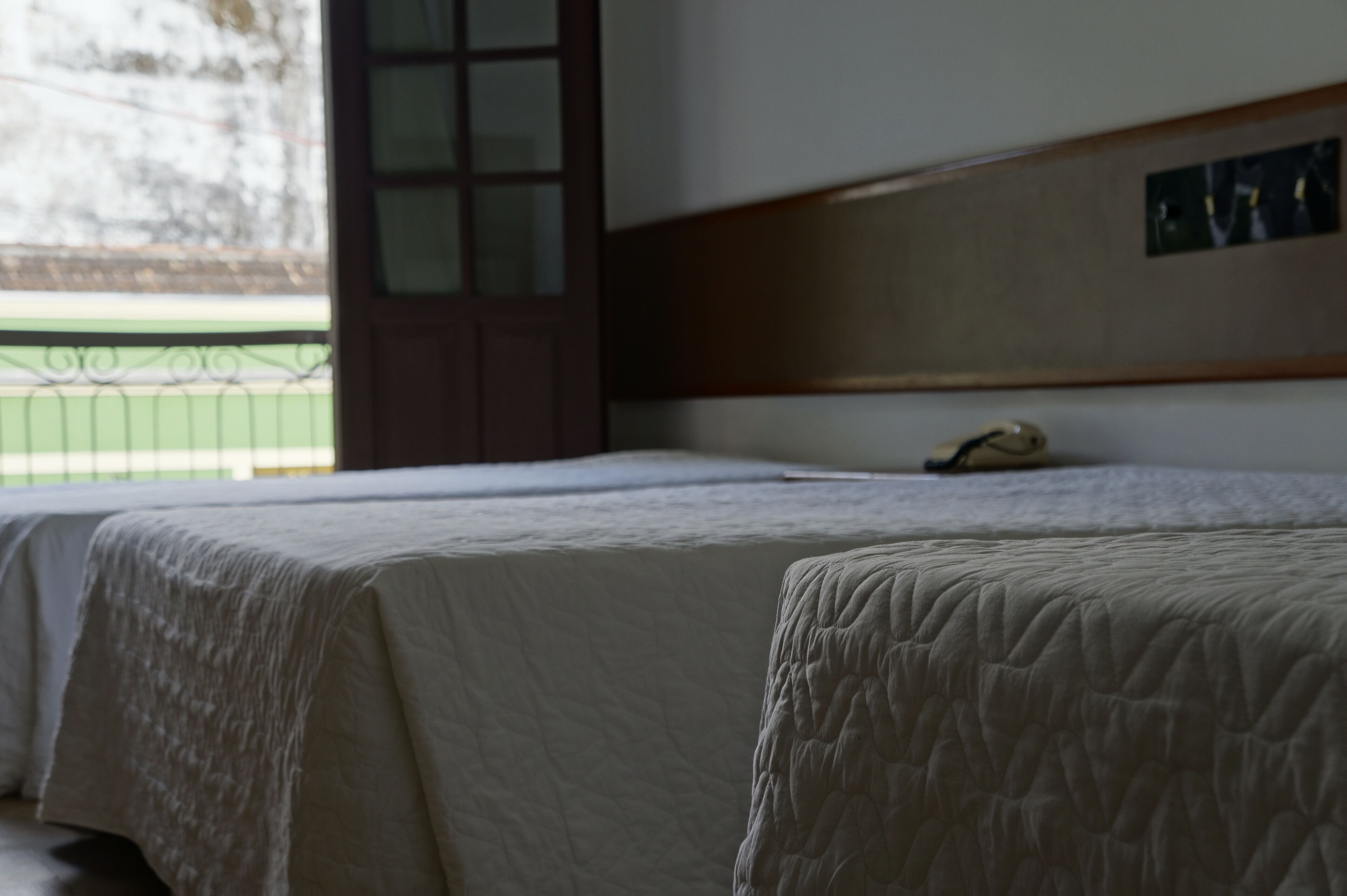 Habitación con dos camas, cabecera de madera, mesita de noche pequeña y ventana con vistas a un espacio exterior verde.