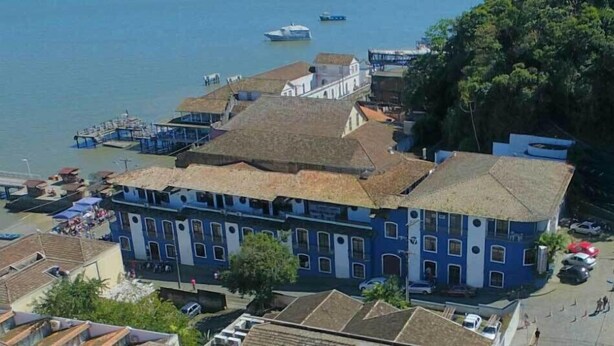 A coastal building with a large bay window, a dock with boats, and a bridge in the background. - Hotel Zibamba (São Francisco do Sul)