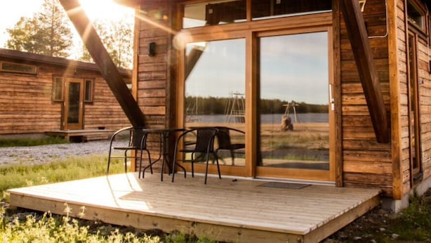 A wooden cabin with a deck, large windows, and a dining area with chairs. - Nordic Lapland Resort (Kalix)