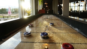 A shuffleboard table with pucks in various positions, a window with a view of the outdoors, and a bar area in the background. - Nordic Lapland Resort (Kalix)