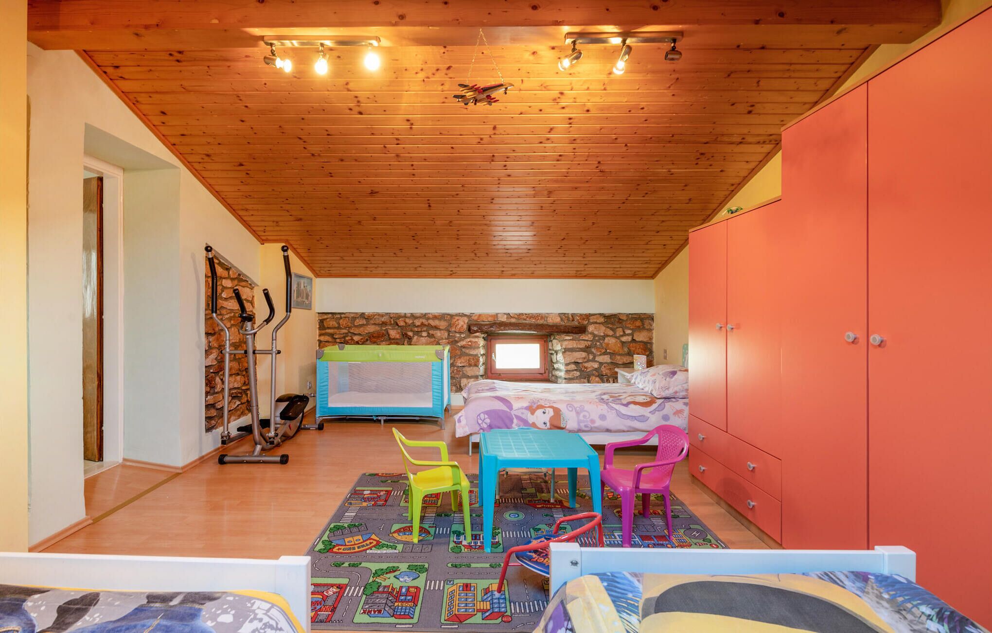 A cozy bedroom with a wooden ceiling, a stone wall, a bed, a desk, and exercise equipment.