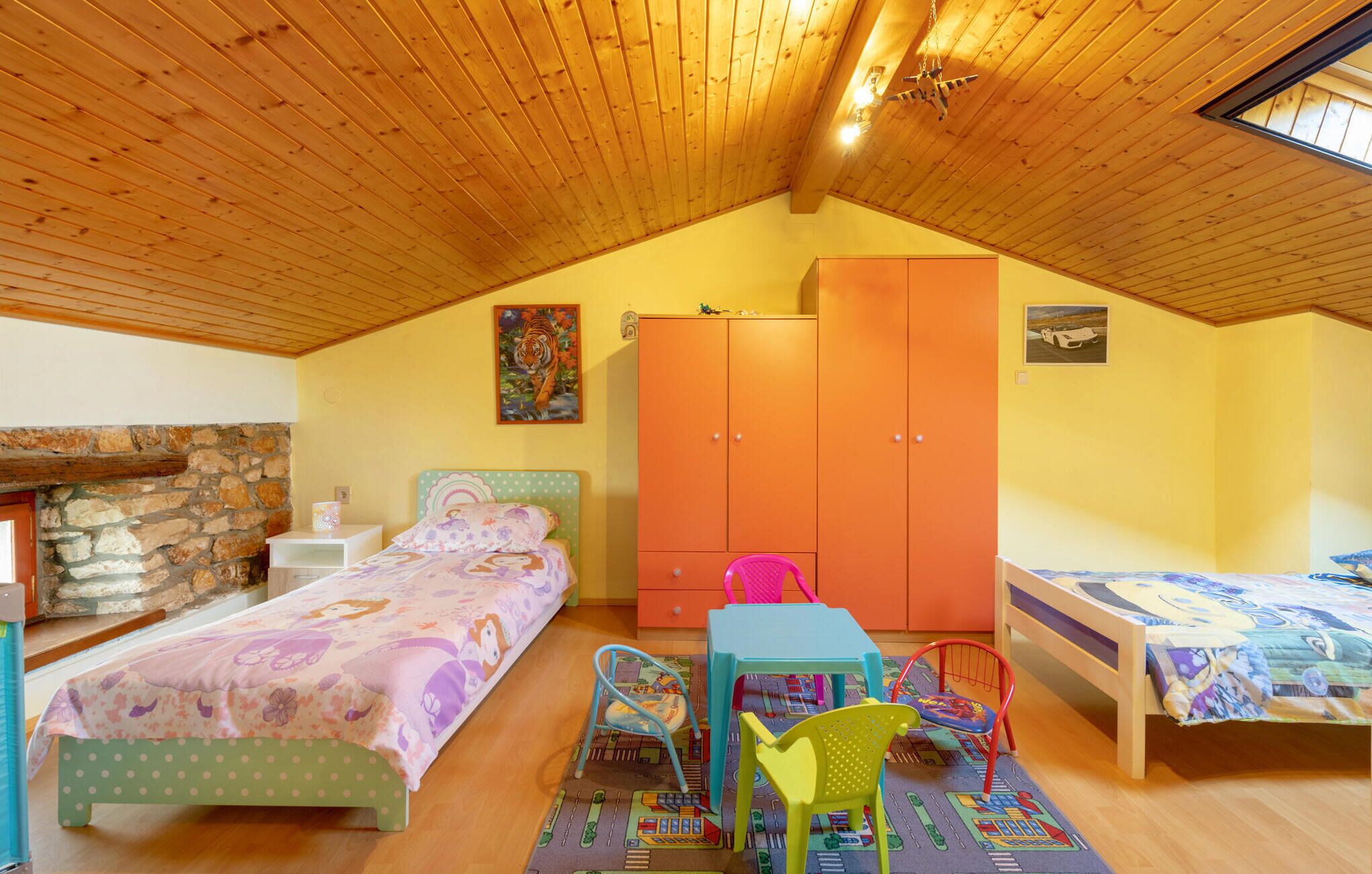 Un dormitorio acogedor en el ático, con techo de madera inclinado, chimenea de piedra, cama con ropa de cama floral, armario y una pequeña mesa con sillas.