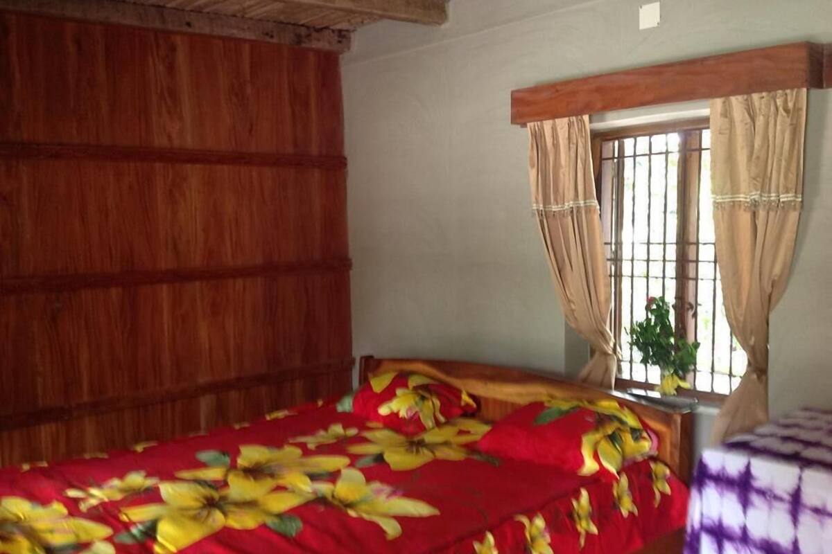 A room with a wooden ceiling, a bed with a floral bedspread, a window with curtains, and a plant on the windowsill.