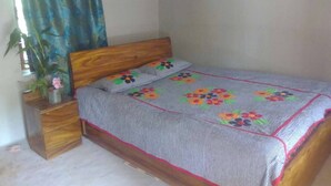 Quarto com cama de madeira, colcha cinza estampada com flores, mesa de cabeceira com um vaso de flores e cortina azul.