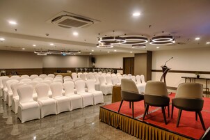 Un salón de conferencias con filas de sillas blancas, un escenario con alfombra roja y modernas luces circulares en el techo.