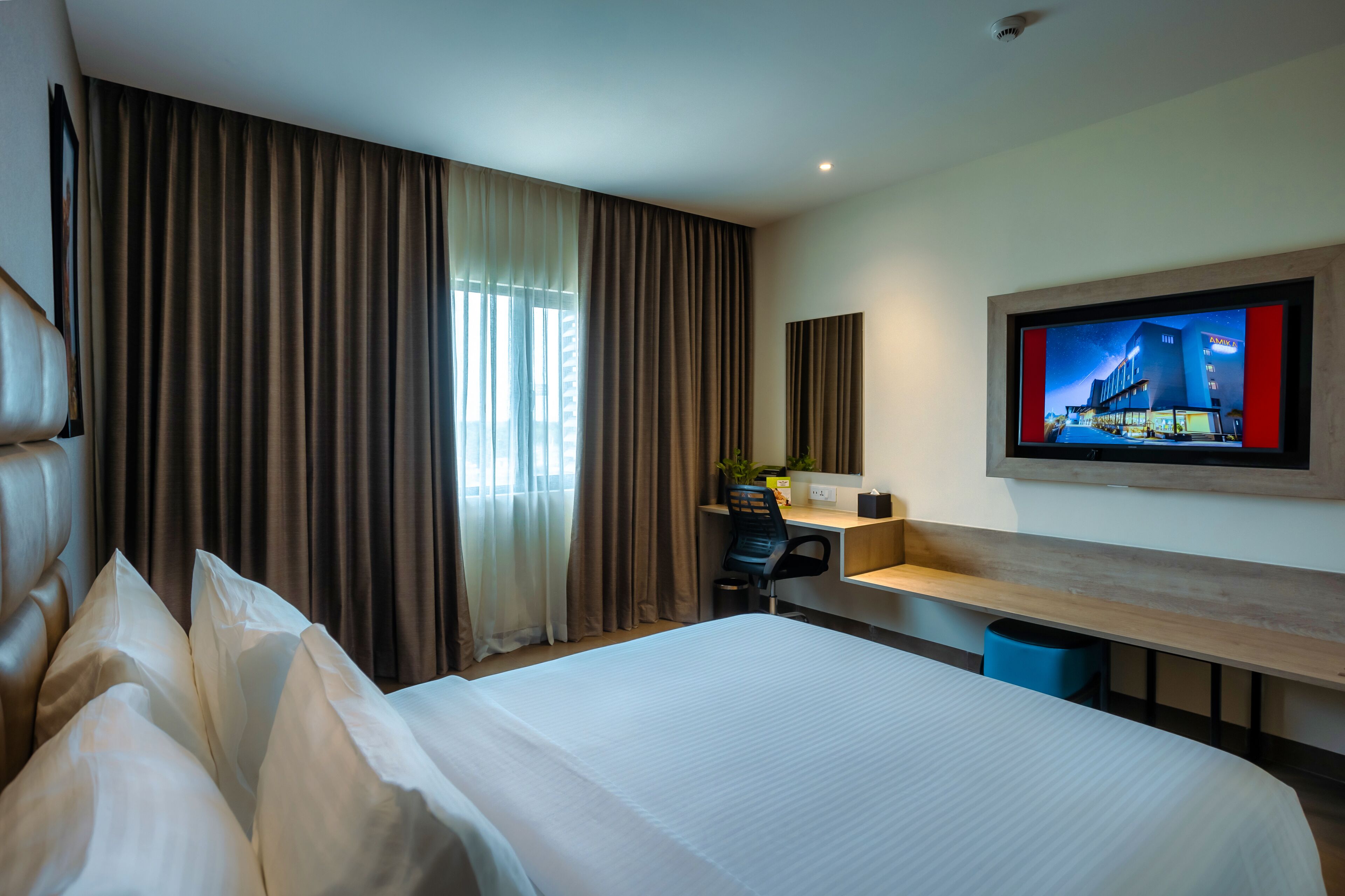 Habitación de hotel con una cama grande, un escritorio con silla, televisor montado en la pared y una ventana con cortinas.