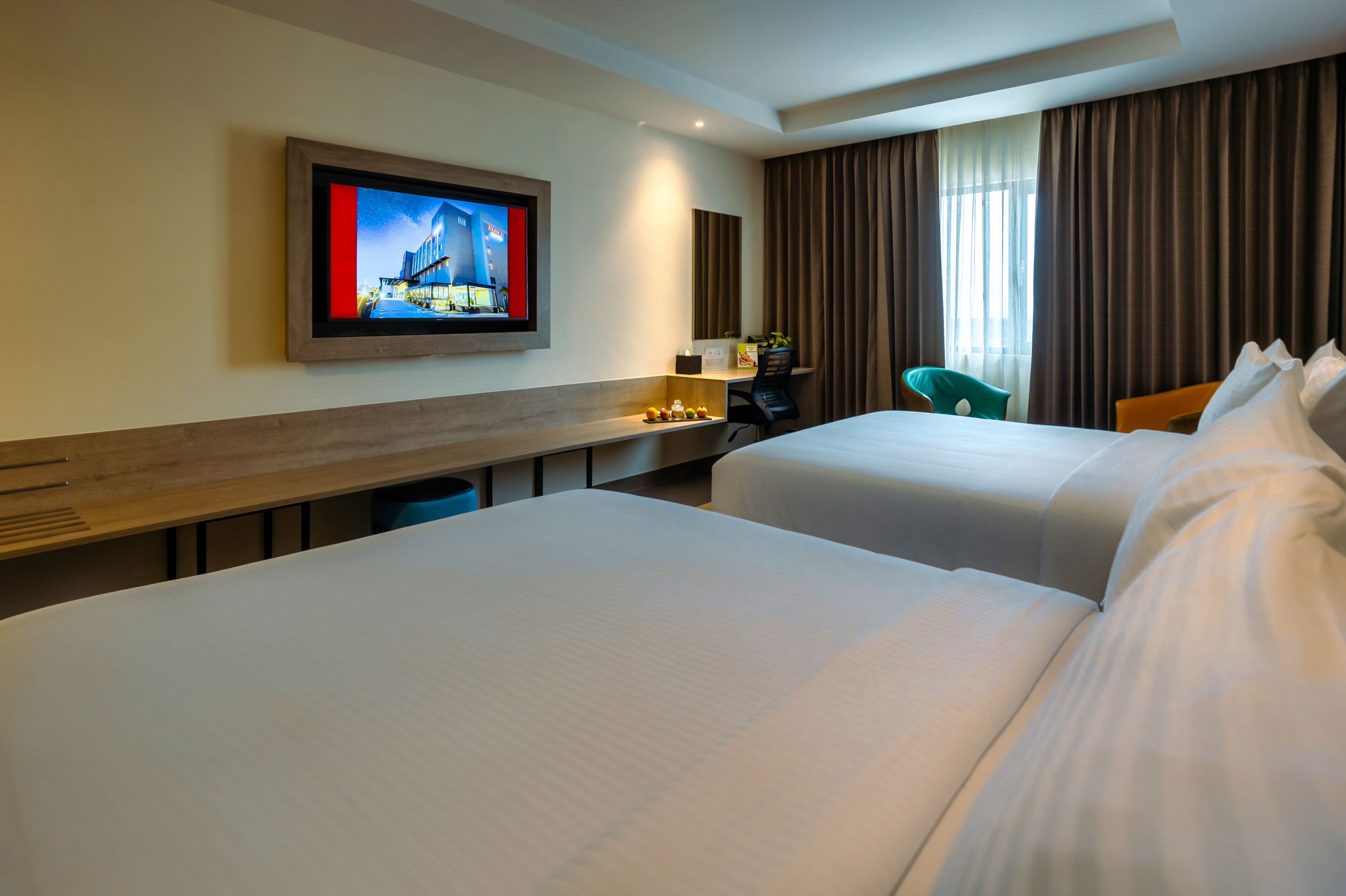 Habitación de hotel con dos camas, un escritorio, una silla, un televisor montado en la pared y una ventana con cortinas.