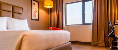 Habitación de hotel con una cama grande, un escritorio, una silla, una ventana y un cuadro en la pared.