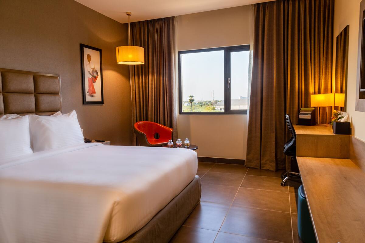 Une chambre d’hôtel avec un grand lit, un bureau, une chaise, une fenêtre avec des rideaux et un tableau au mur.