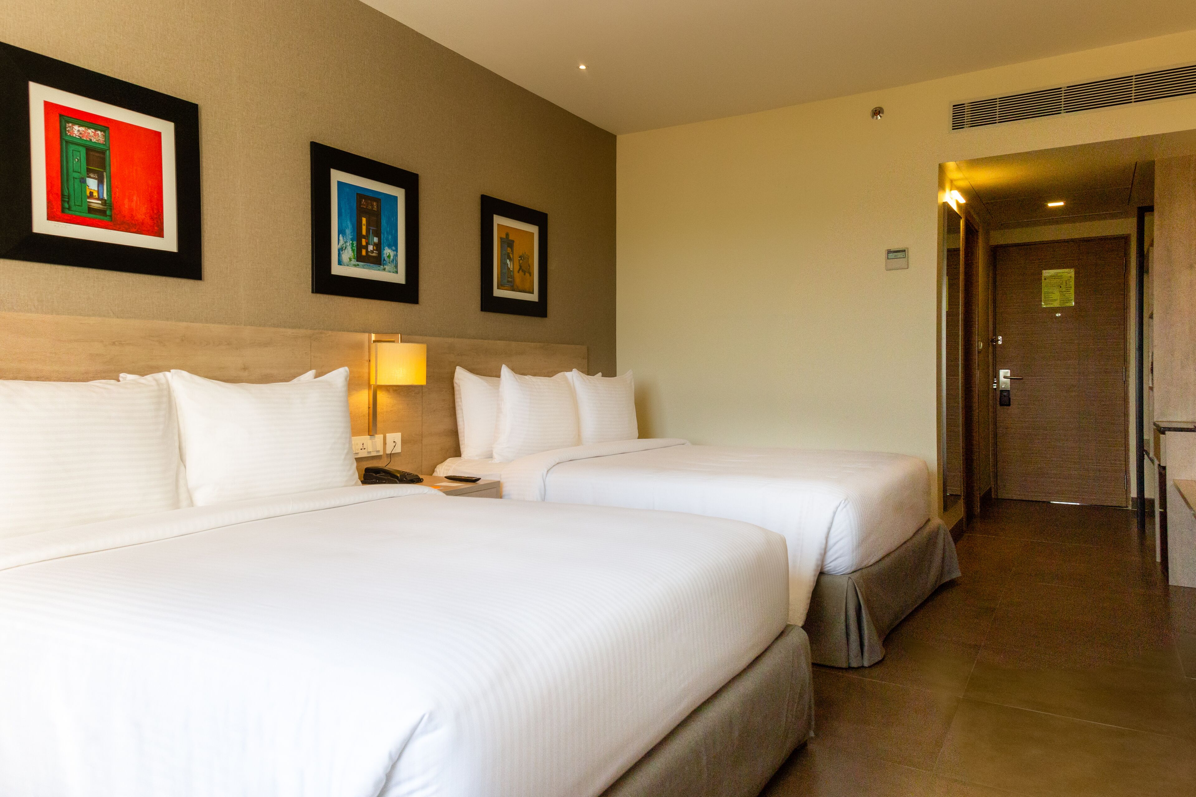 A hotel room with two beds, a nightstand, a lamp, and framed artwork on the wall.