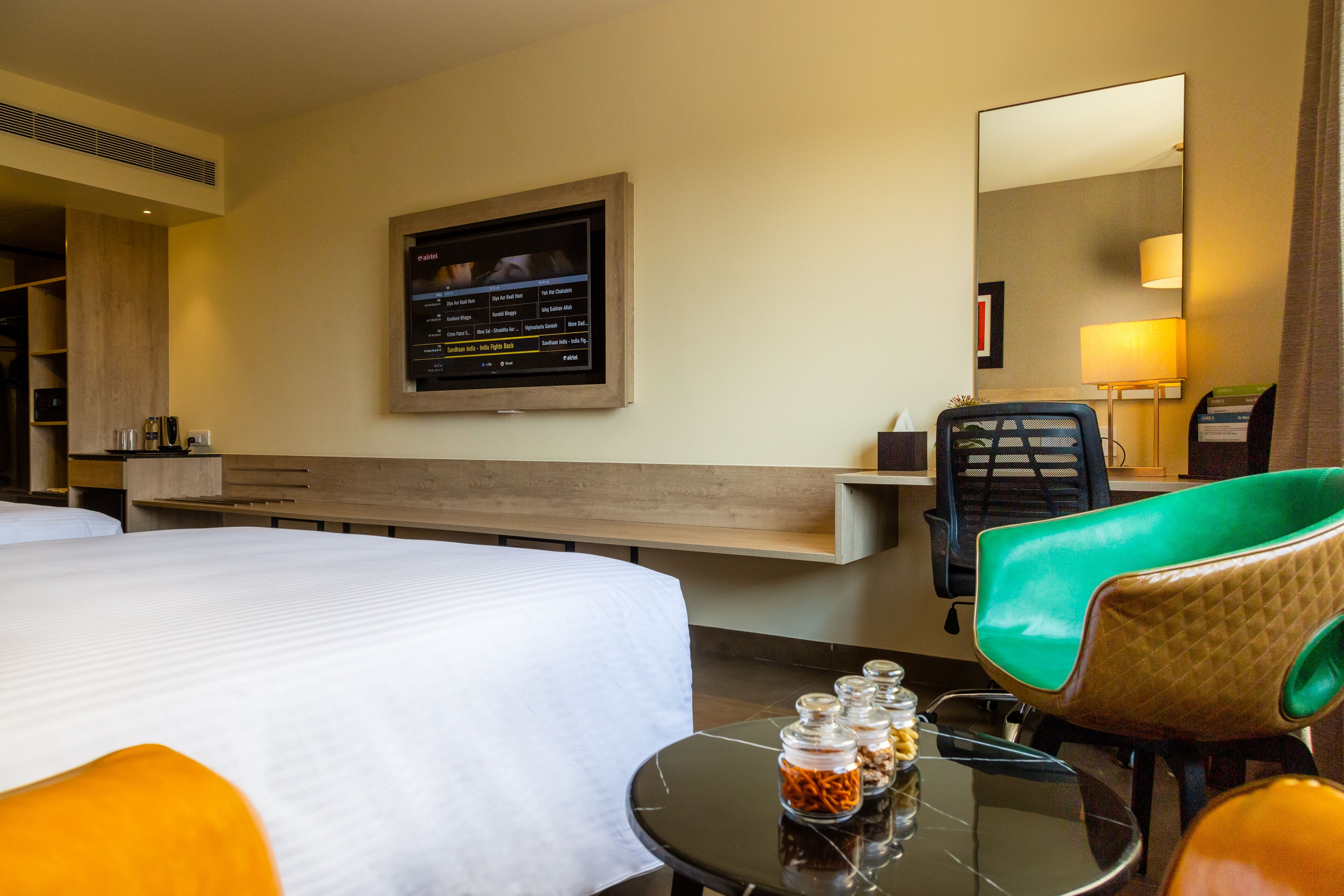 A hotel room with a bed, a television mounted on the wall, a desk with a chair, and a small table with jars of snacks.