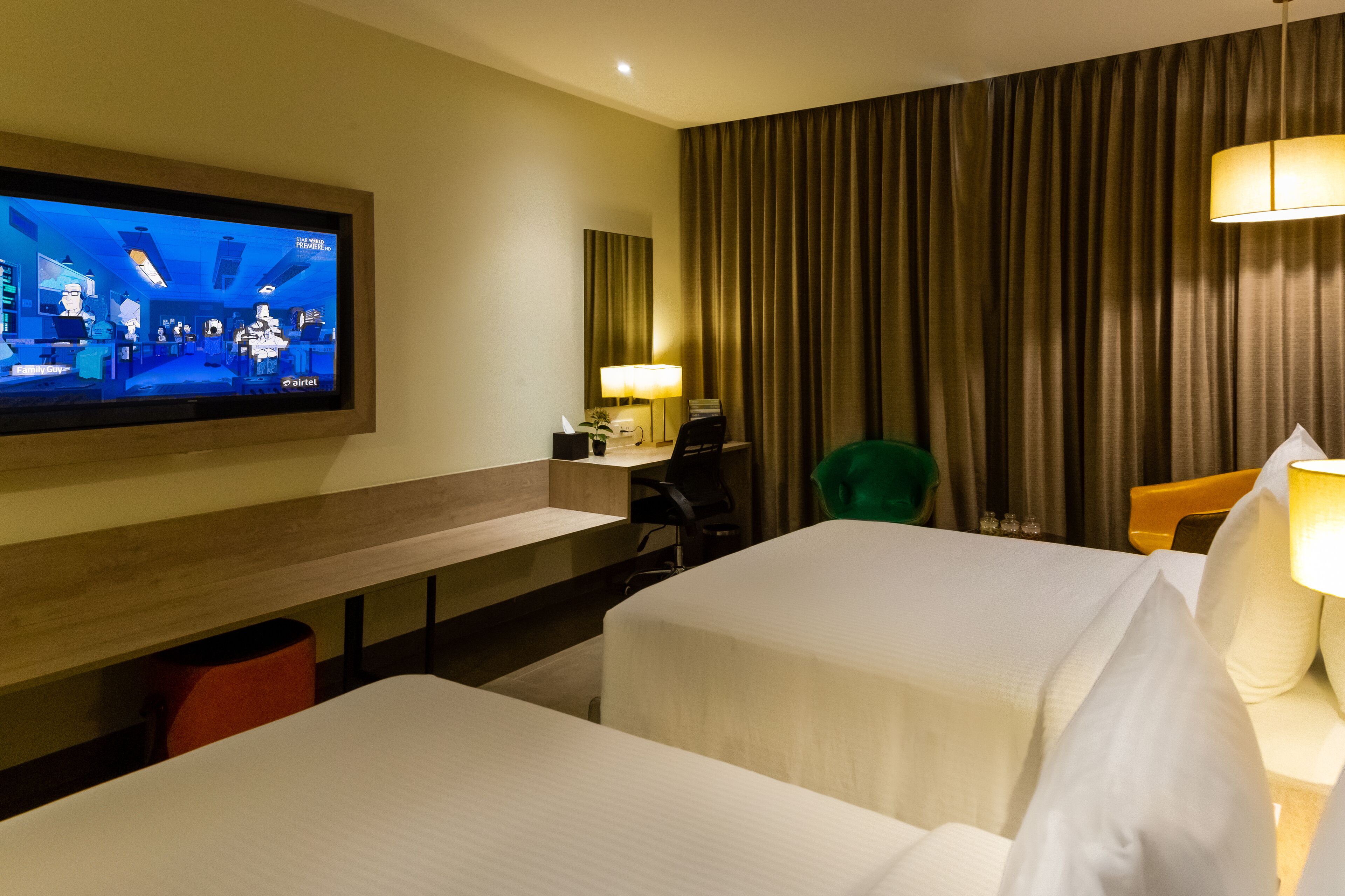 A hotel room with two beds, a desk, a chair, a TV, and a lamp.