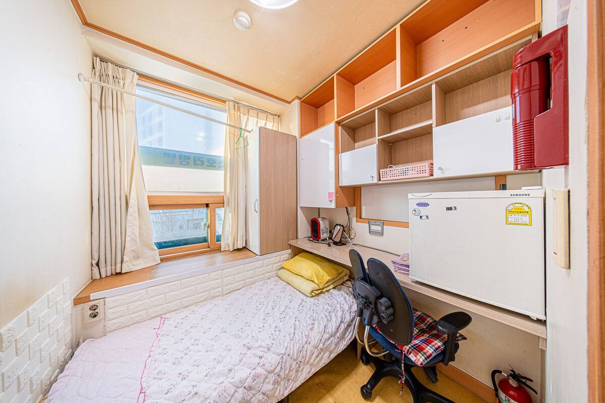 A compact room with a single bed, a desk with a chair, a small window with curtains, and a white refrigerator.