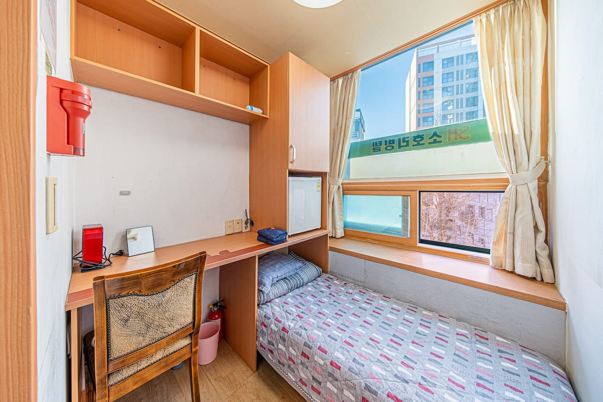 A compact room with a bunk bed, a desk with a chair, a small window with curtains, and a view of buildings outside.