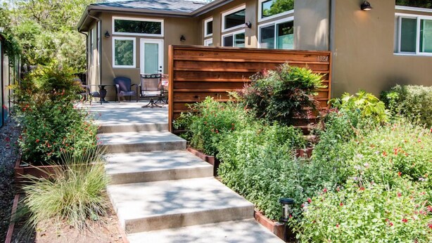 A modern house with a wooden deck, a staircase leading to the front door, and a garden with various plants and flowers.