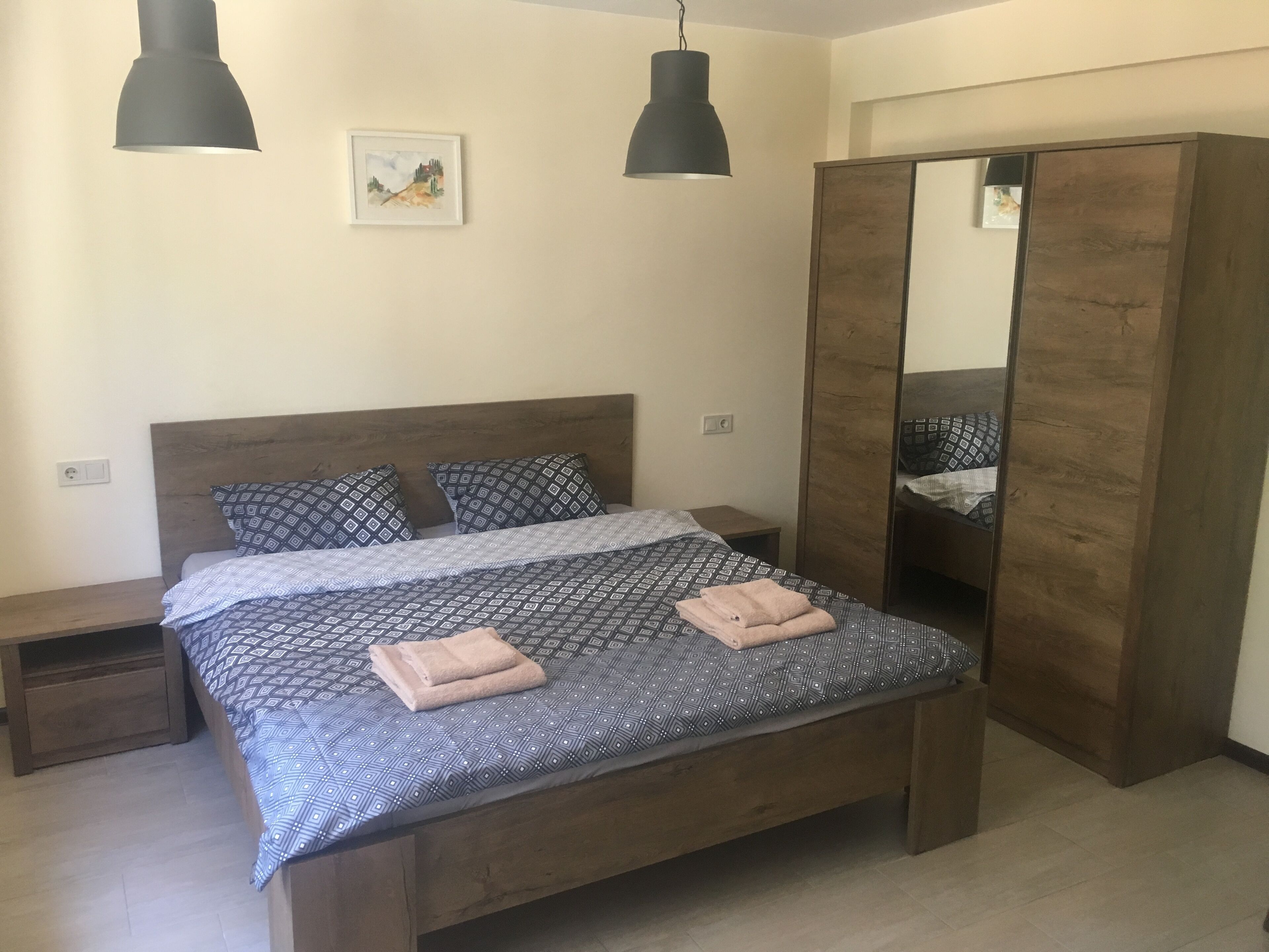 A neatly made bed with patterned bedding, two bedside tables, a large mirror, and two pendant lights.