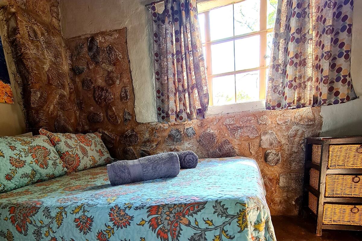 Une chambre rustique avec un mur en pierre, un lit recouvert d’une literie à motifs floraux et une fenêtre ornée de rideaux à dessins.