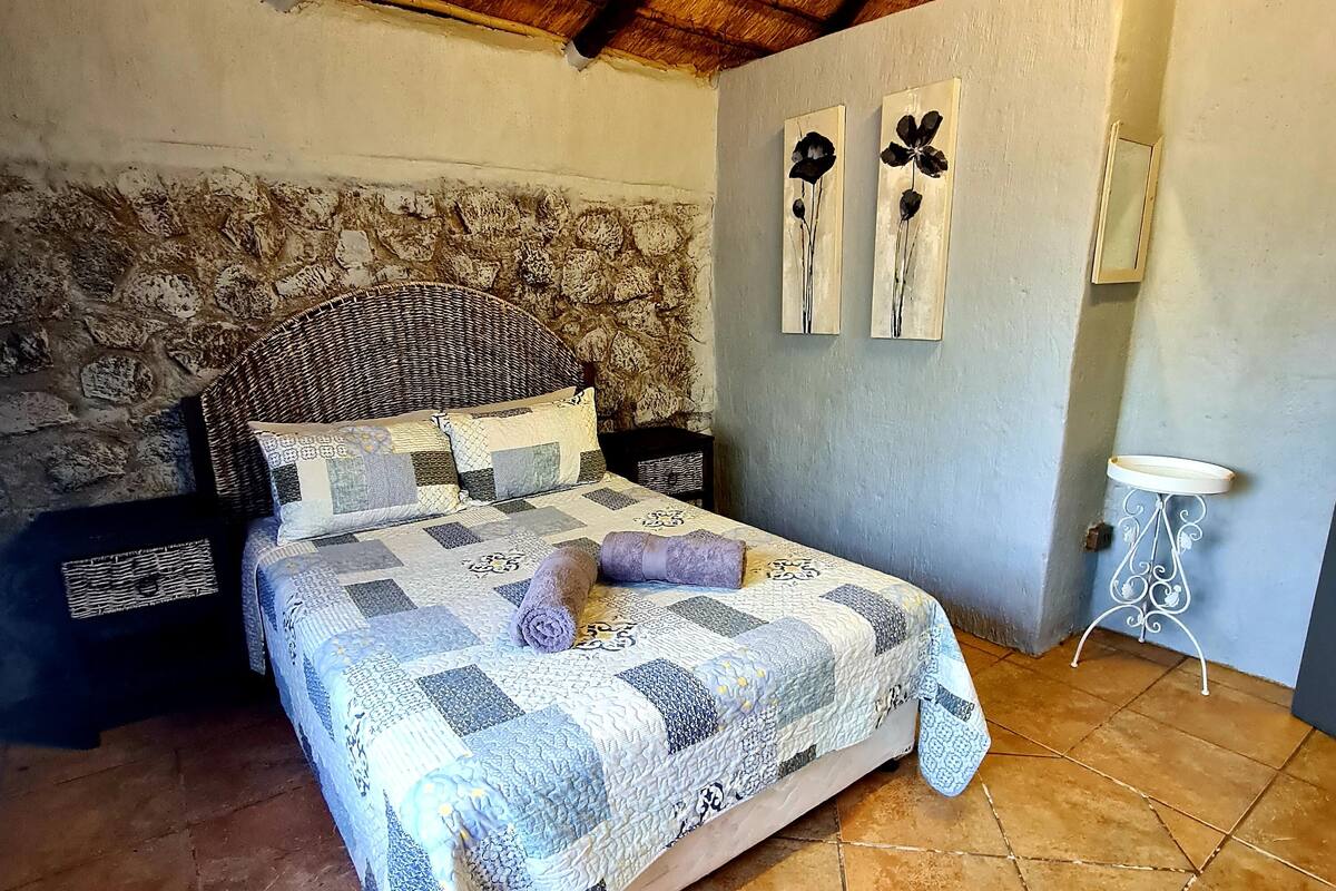 Une chambre avec un mur en pierre, un lit recouvert d’une courtepointe, deux tableaux représentant des fleurs accrochés au mur et une petite table blanche.