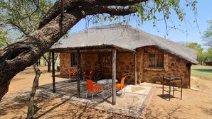 A stone building with a thatched roof, a covered outdoor seating area with orange chairs, and a dirt ground. - Uitvlugt Resort (Bela-Bela)