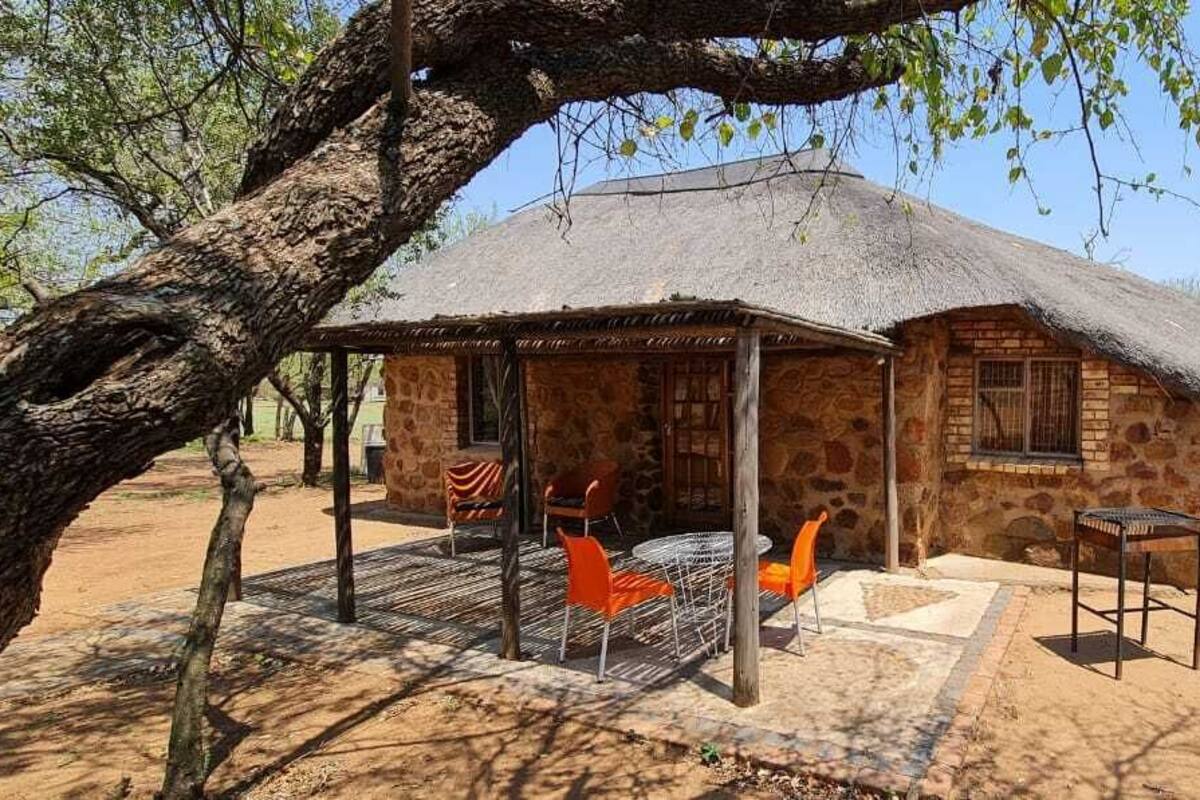 Un bâtiment en pierre avec un toit de chaume, un espace extérieur abrité avec des chaises orange, et un sol en terre battue.
