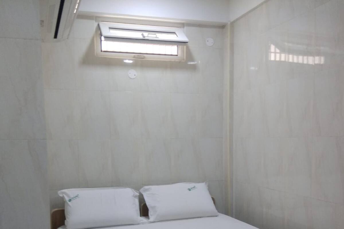 Une petite chambre avec un lit simple, des murs blancs, une fenêtre fermée par un store et un lit recouvert d’une literie blanche et d’une couverture colorée.