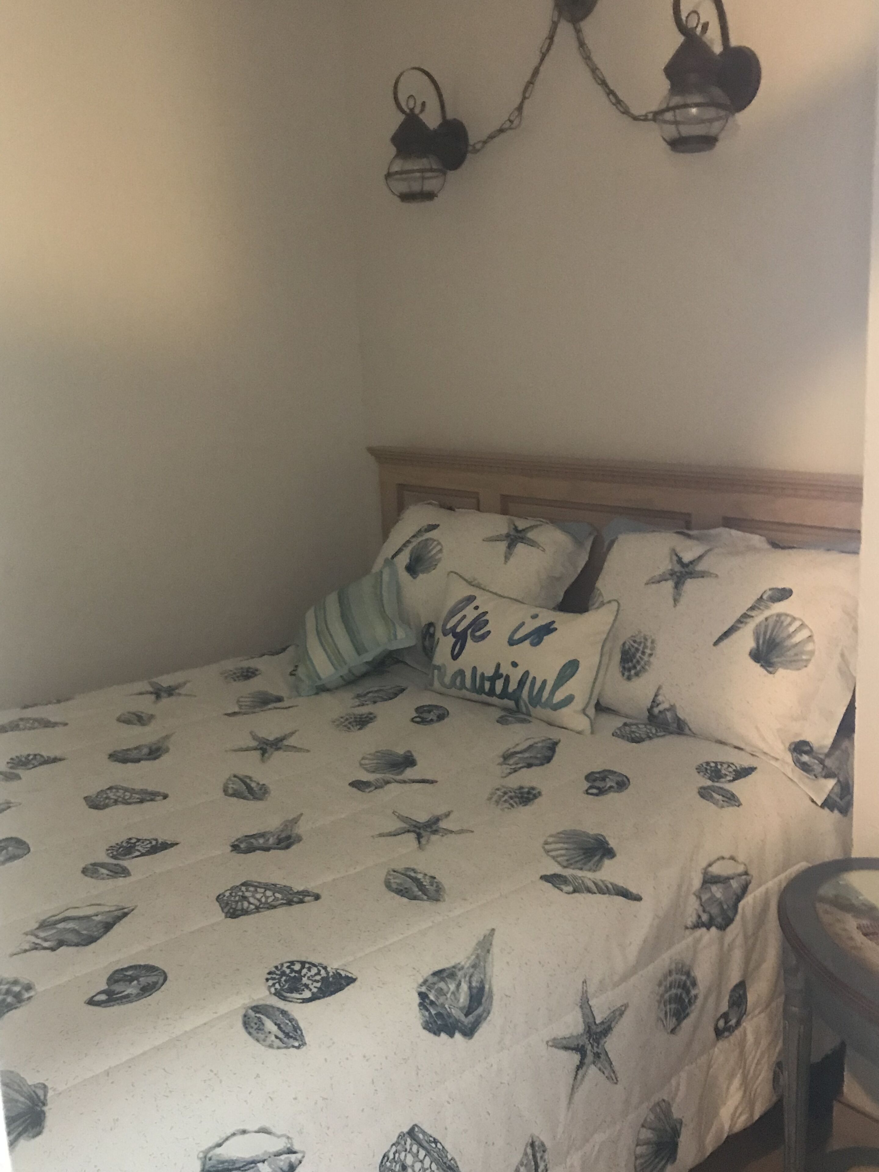A bed with a seashell and starfish pattern, a pillow with 'Life is Beautiful' text, and a small table beside the bed.