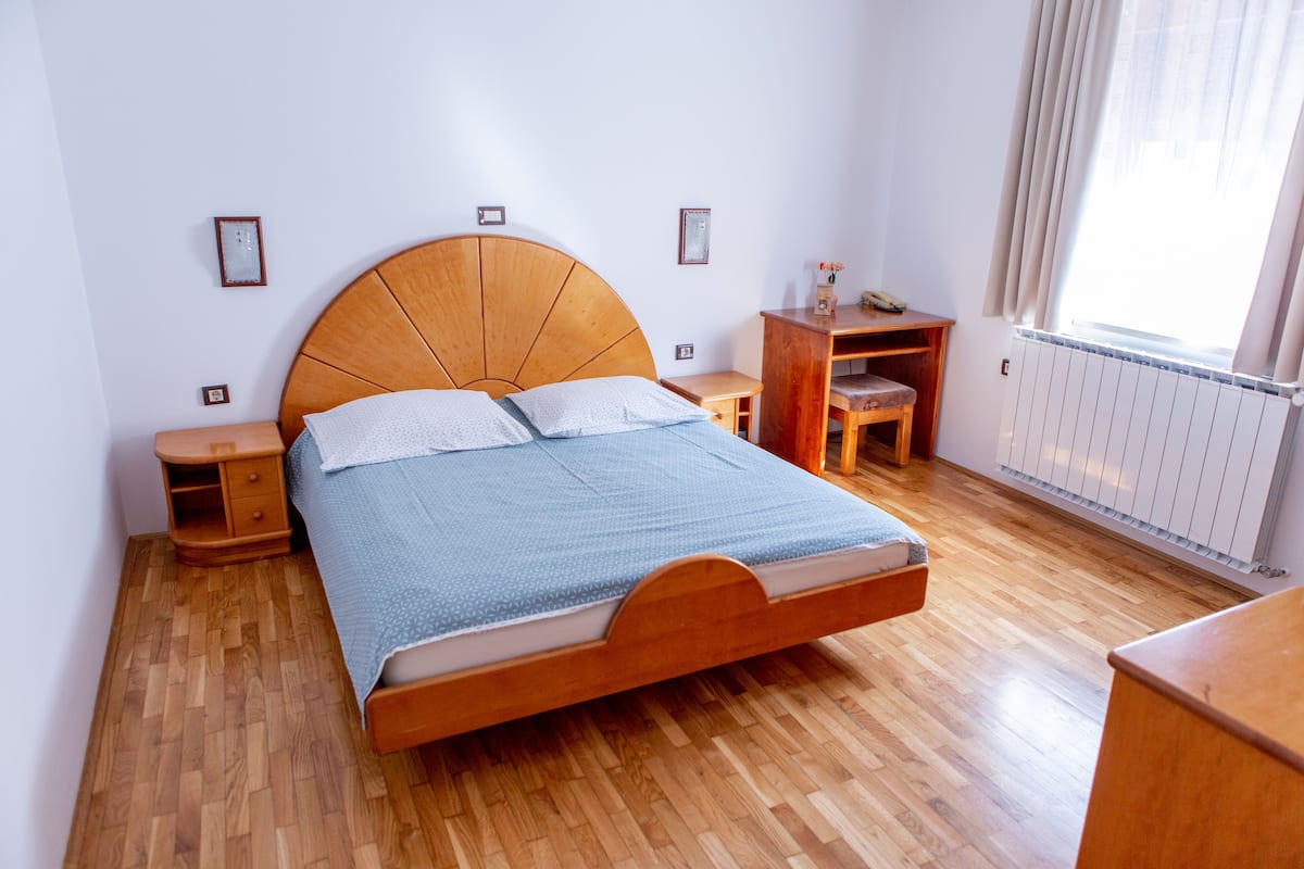 A bedroom with a wooden bed, two bedside tables, a nightstand, a wooden dresser, and a window with curtains.