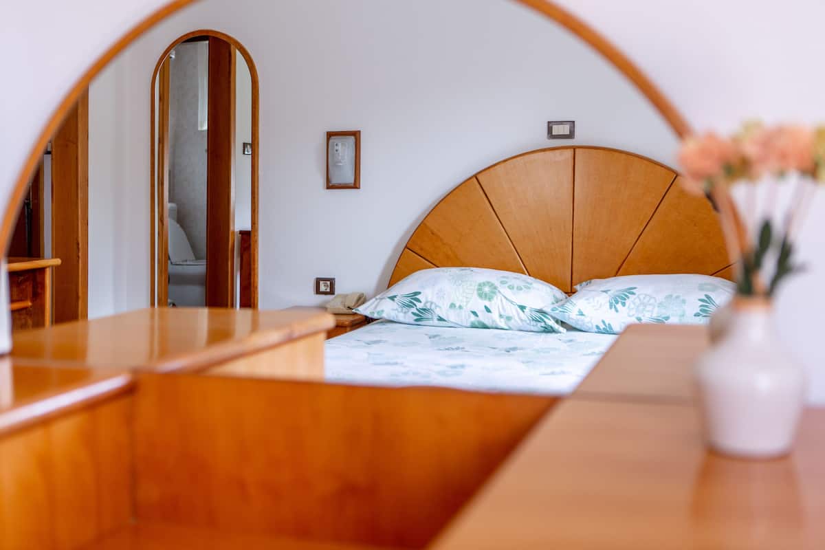 A bedroom with a wooden headboard, a bed with floral bedding, a bedside table, a mirror, and a vase with flowers.