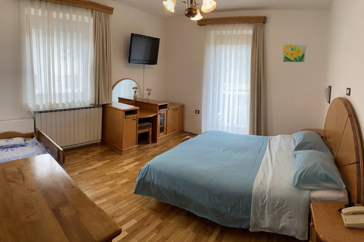 A bedroom with a wooden bed, a desk with a chair, a small vanity, and a television on the wall.