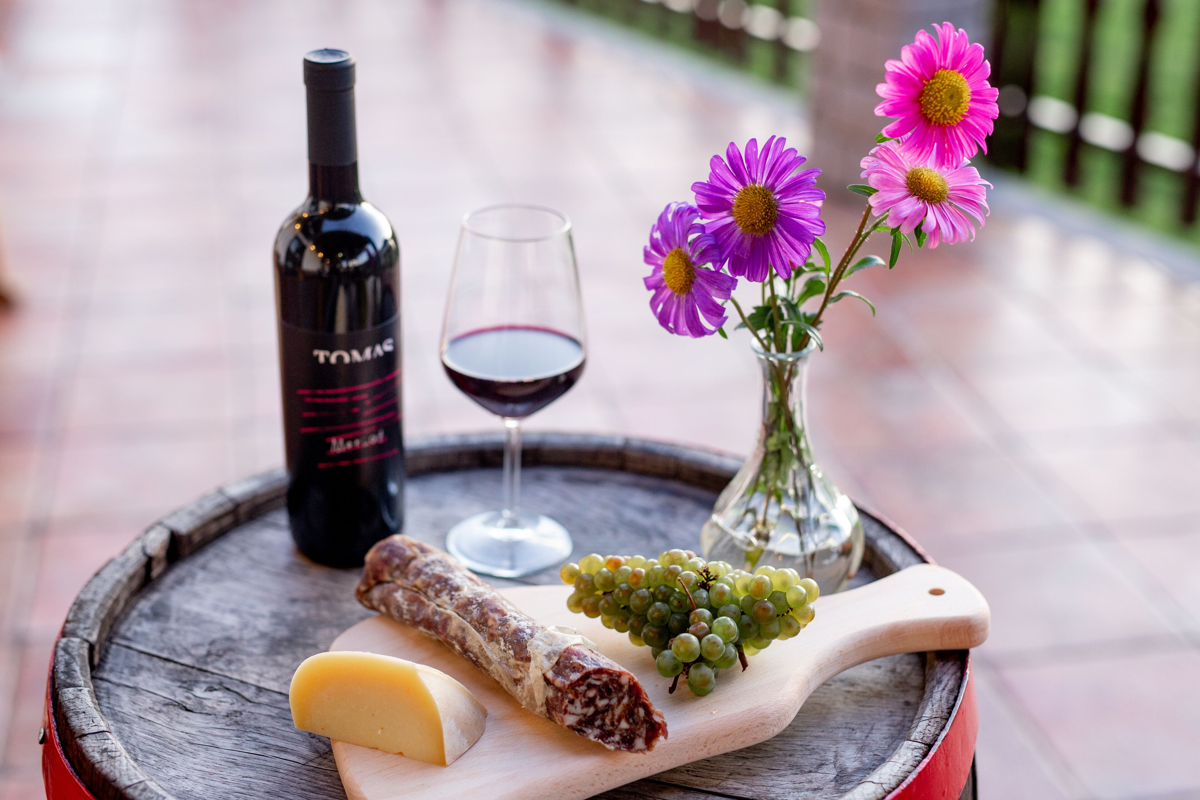 A wooden barrel table with a bottle of wine, a glass of wine, a cheese wheel, a piece of cured meat, a bunch of grapes, and a vase with flowers.