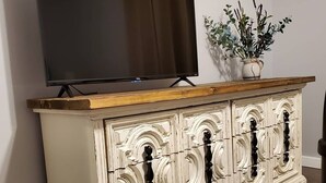 Un mueble blanco y ornamentado con un televisor encima, una planta pequeña y una encimera de madera.