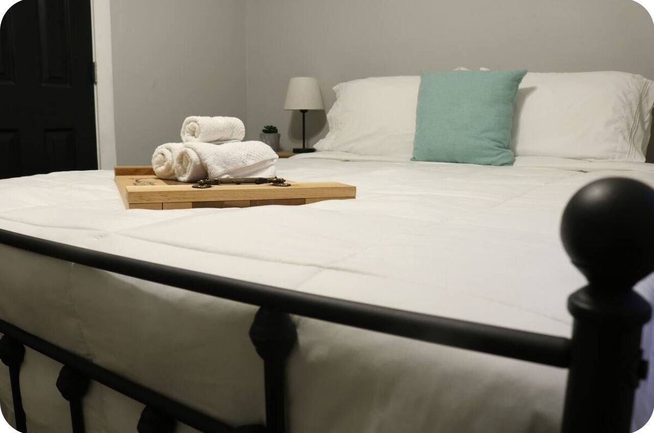 Een netjes opgemaakt bed met witte beddengoed, een blauwgroen kussen en een dienblad met opgevouwen handdoeken.