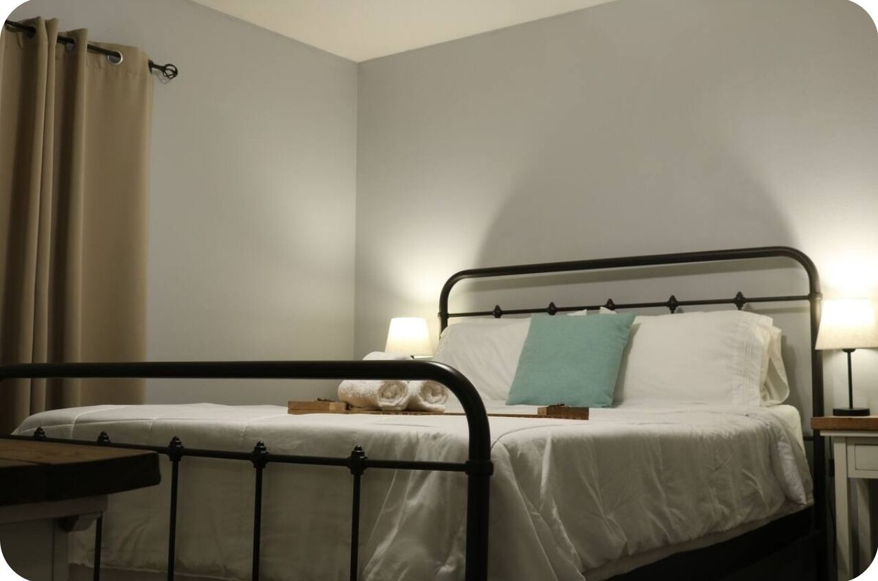 Una cama individual con estructura metálica, ropa de cama blanca y un cojín verde azulado. Hay dos lámparas de noche de madera.