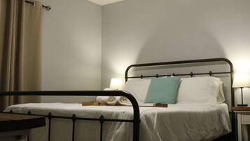 Una cama individual con estructura metálica, ropa de cama blanca y un cojín verde azulado. Hay dos lámparas de noche de madera.