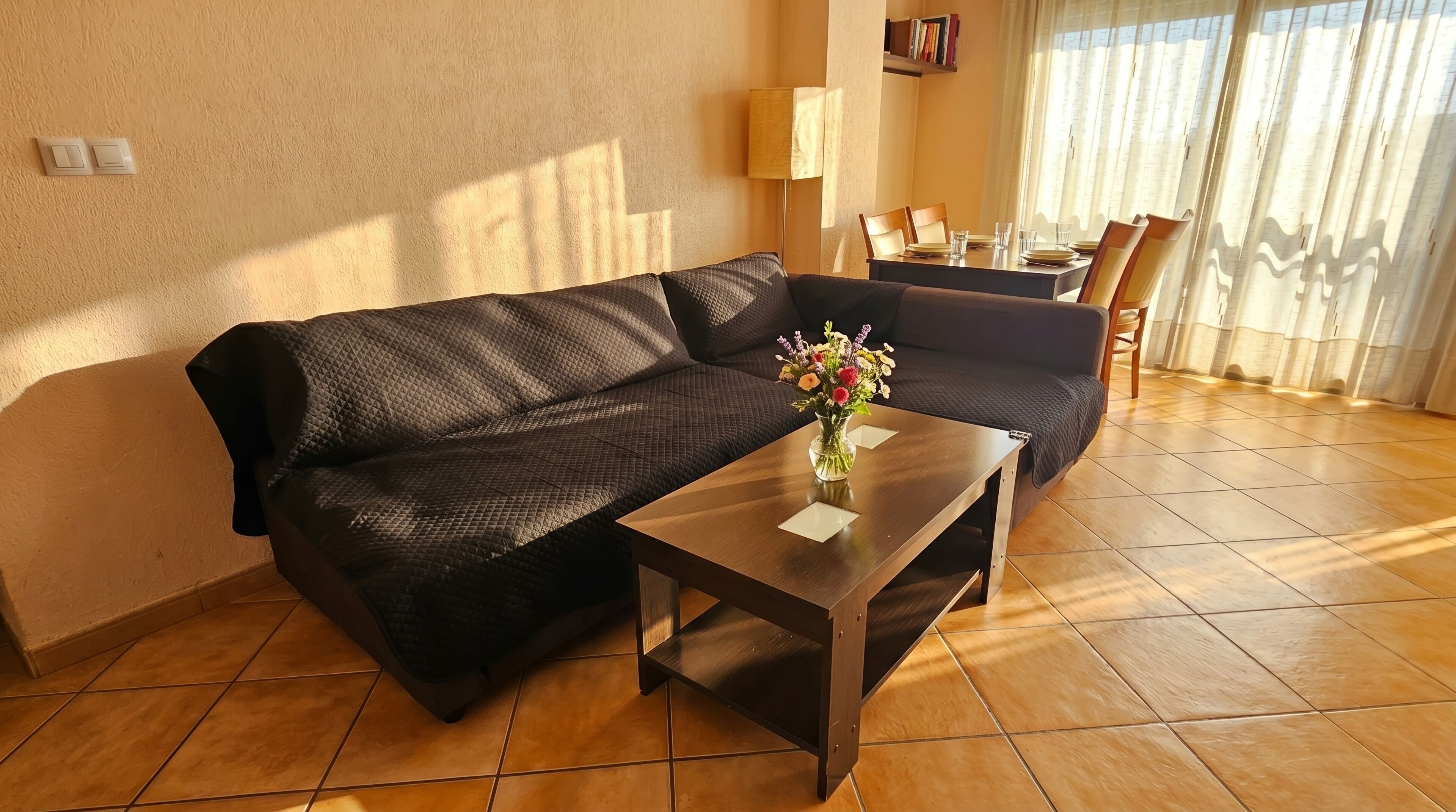 Un salon avec un canapé marron foncé, une table basse en bois ornée d’un vase de fleurs et un coin repas avec une table dressée pour quatre personnes.