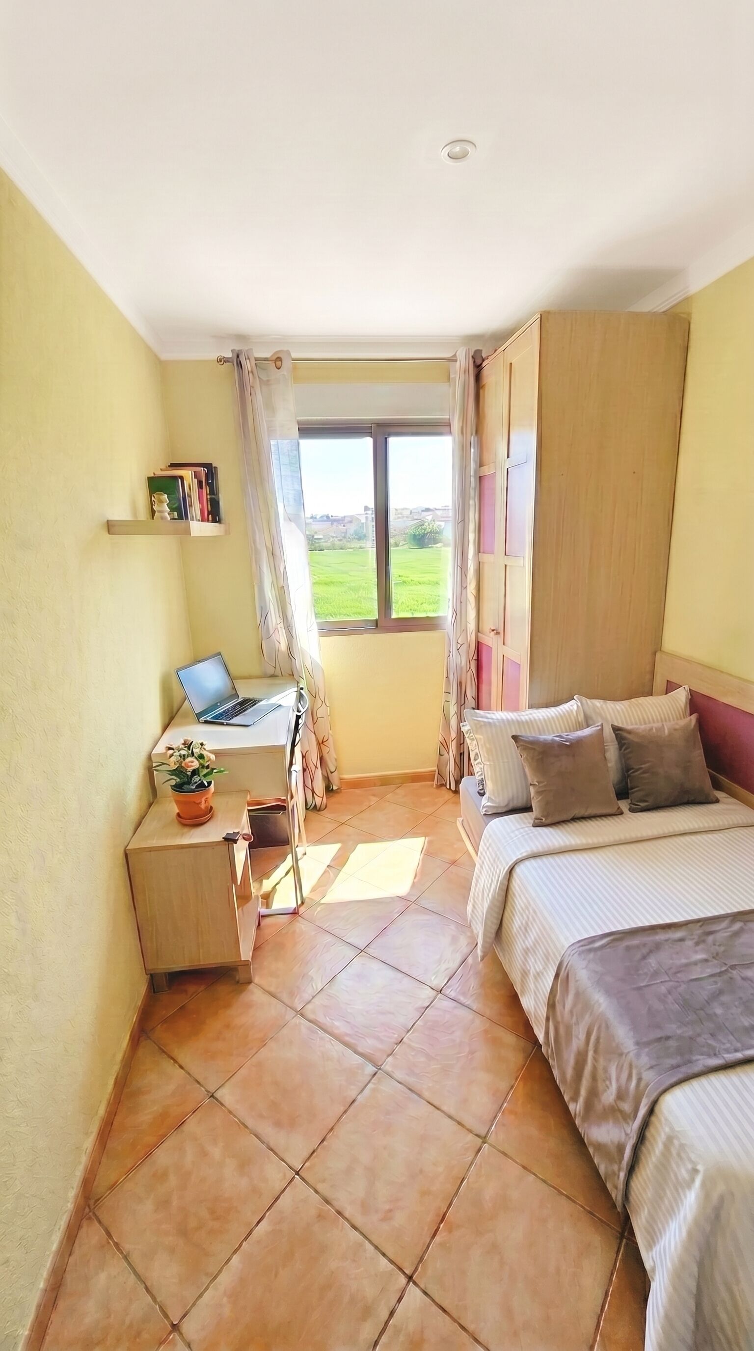 A bedroom with a bed, a desk with a laptop, a window with curtains, and a view of greenery outside.