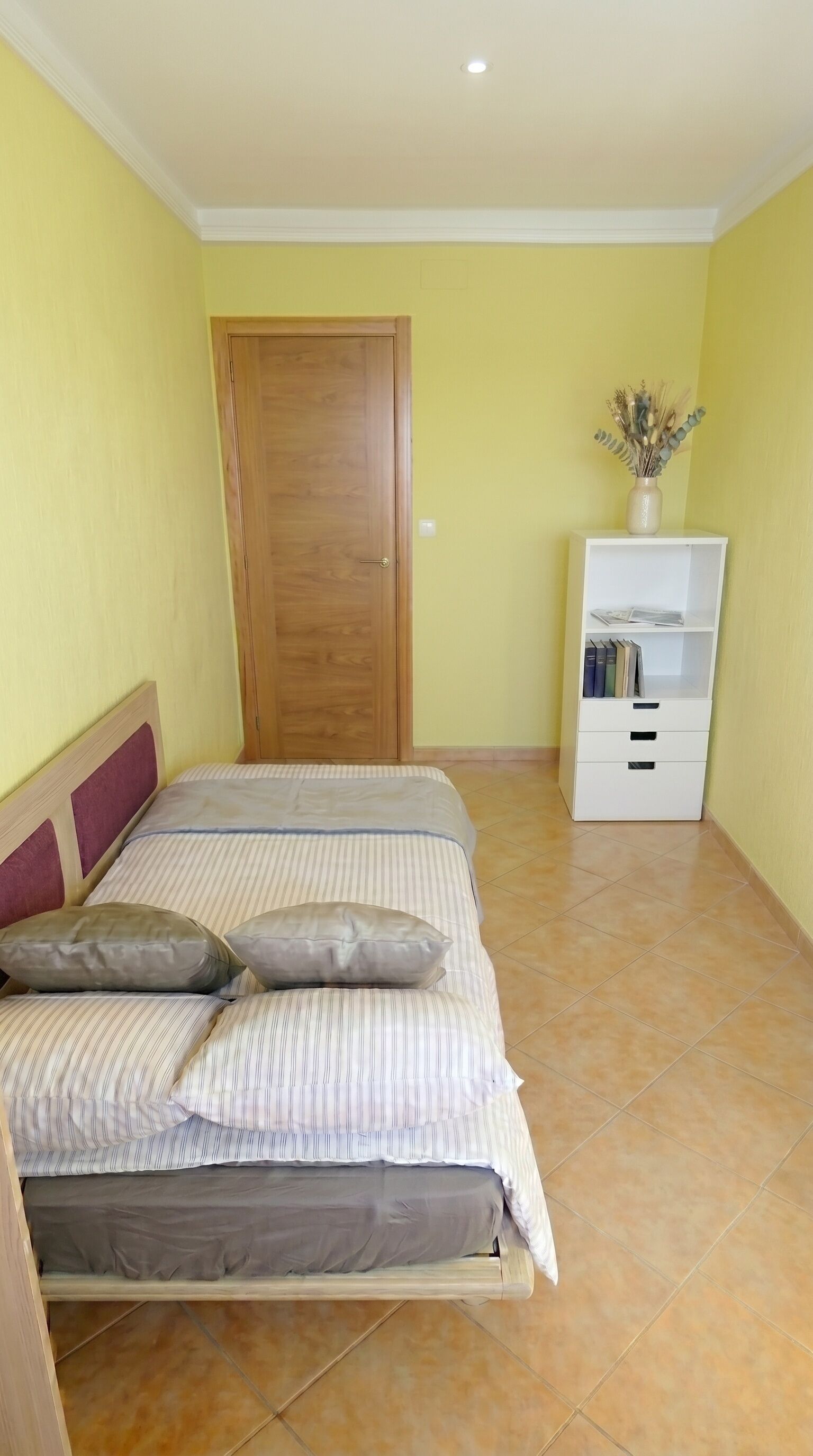 Une chambre à coucher avec un lit, une porte en bois, une armoire blanche et un vase contenant des plantes séchées.