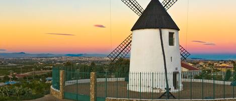 Eine Windmühle mit weißem Korpus und schwarzem Dach, umgeben von einem grünen Zaun und einer Steinmauer, vor einem Sonnenuntergangshimmel.