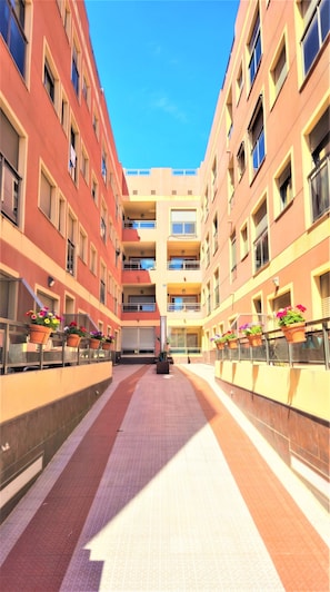 A corridor with a tiled floor, flanked by buildings with balconies and potted plants.