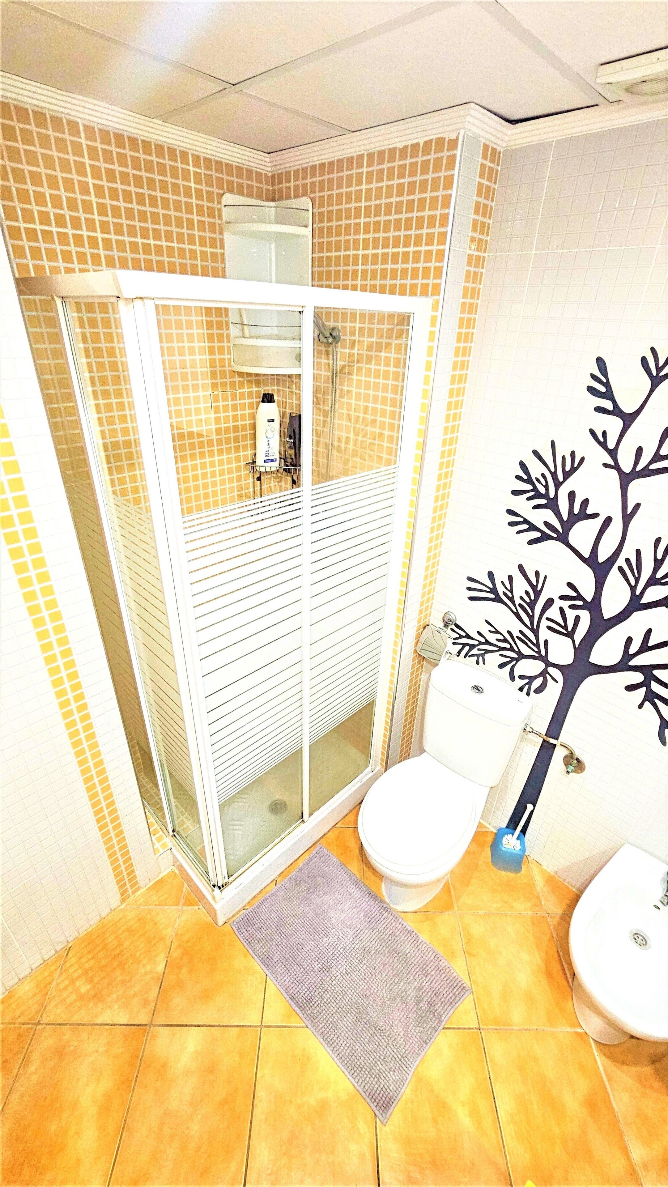 Un baño con cabina de ducha, dos inodoros, un lavabo y un mural decorativo con motivos vegetales.
