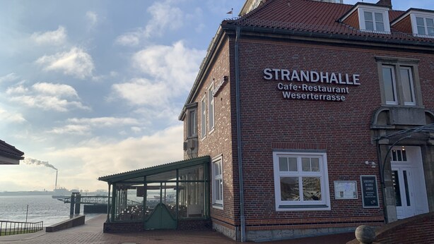 A brick building with a sign reading "STRANDHALLE Cafe Restaurant Weserterasse" and a covered outdoor seating area. - Apartment Seeweg - your time out in the north (Beverstedt)