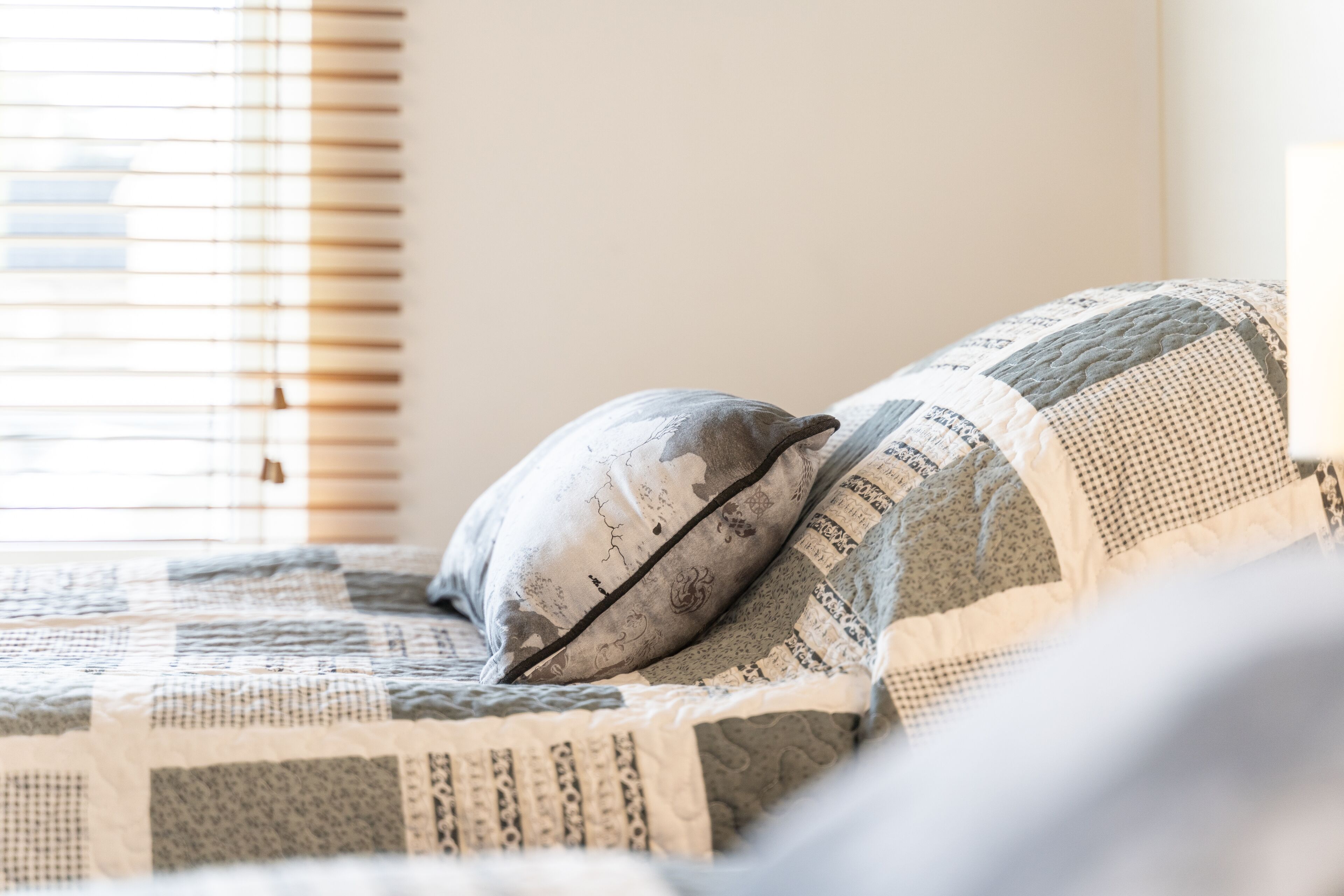 A bed with a quilted blanket and pillows, a window with blinds, and a wall.