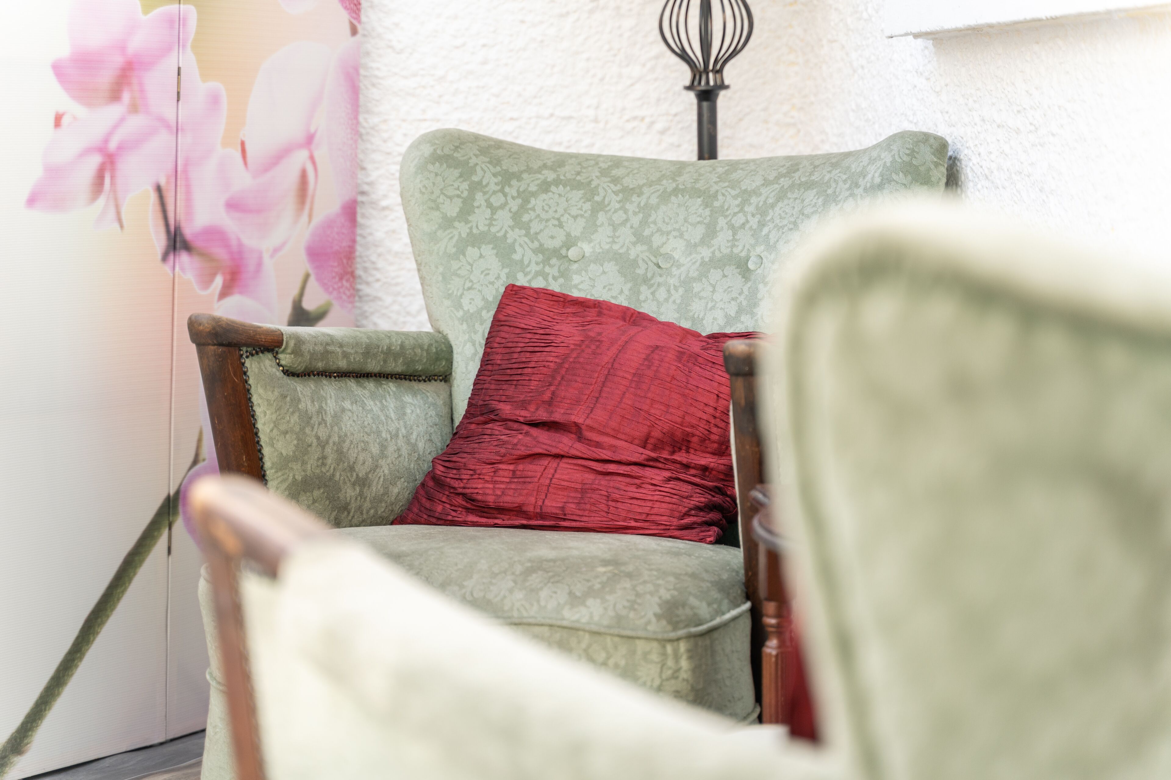 A chair with a floral pattern and a red cushion, positioned in front of a wall with a floral painting.