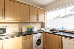 A kitchen with wooden cabinets, a washing machine, a microwave, and a window with a view of buildings. - 3 Bed Quaint House Town Center (Wexford)